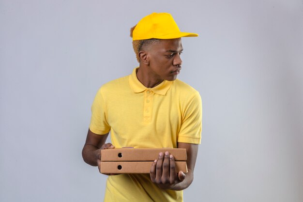 Homme afro-américain de livraison mécontent en polo jaune et casquette tenant des boîtes de pizza à côté avec froncement de sourcils debout
