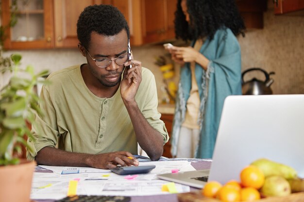 Homme afro-américain malheureux grave ayant une conversation téléphonique lors du calcul du budget familial dans la cuisine