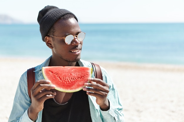 Homme afro-américain à la mode en chapeau noir et lunettes de soleil se sentant heureux et insouciant tenant une grosse tranche de riche pastèque mûre rouge tout en passant la journée d'été à l'extérieur sur la plage urbaine