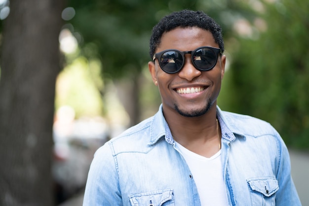 Homme afro-américain portant des lunettes de soleil et souriant tout en se tenant à l'extérieur dans la rue.