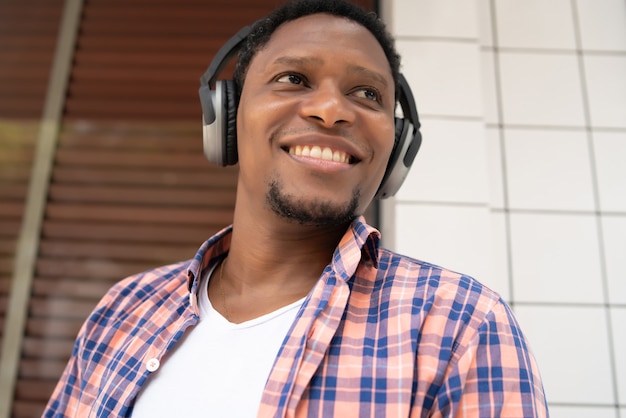 Homme afro-américain souriant et écoutant de la musique avec des écouteurs tout en se tenant à l'extérieur dans la rue