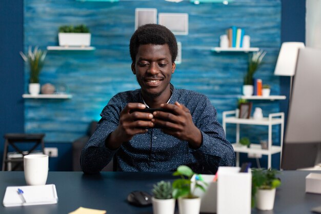 Homme afro-américain souriant tout en utilisant un smartphone au bureau pour vérifier le travail à distance sur les réseaux sociaux...