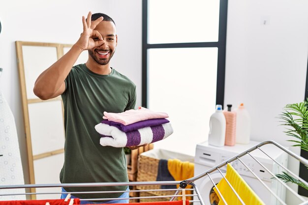 Homme afro-américain tenant le linge plié de la corde à linge souriant heureux faisant signe ok avec la main sur les yeux regardant à travers les doigts