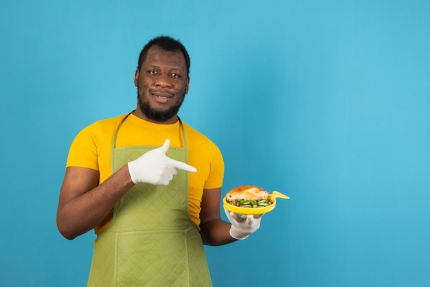 Homme afro-américain tenant avec repas sur mur bleu heureux pointant avec la main et le doigt.