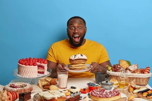 L'homme afro-américain tient une assiette avec des crêpes crémeuses et des baies, garde la bouche ouverte, a surpris l'expression, vêtu d'un t-shirt jaune décontracté