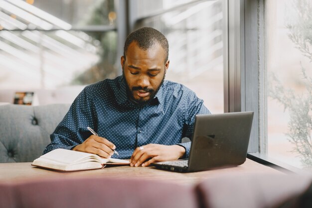 Homme afro-américain travaillant derrière un ordinateur portable et écrivant dans un cahier. Homme à la barbe assis dans un café.