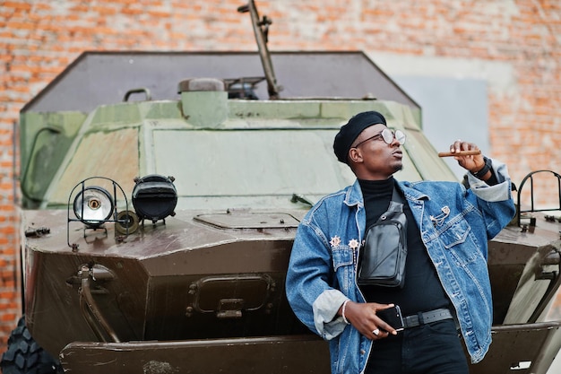 Photo gratuite homme afro-américain en veste de jeans béret et lunettes avec cigare posé contre un véhicule blindé militaire btr
