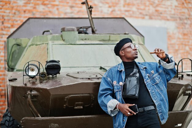 Homme afro-américain en veste de jeans béret et lunettes avec cigare posé contre un véhicule blindé militaire btr