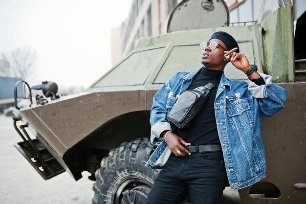 Homme afro-américain en veste de jeans béret et lunettes fumant un cigare et posé contre un véhicule blindé militaire btr