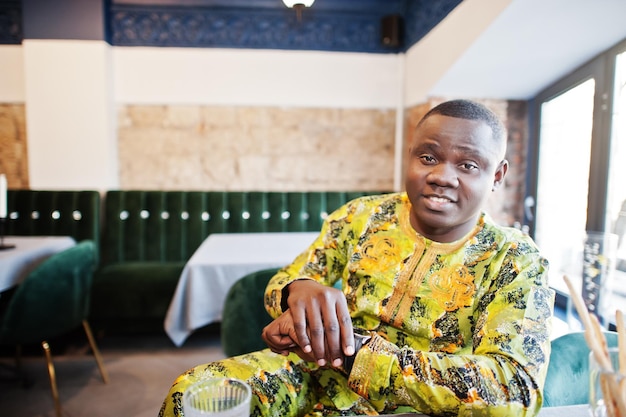 Homme afro amical en vêtements traditionnels avec casquette assis au restaurant et regardant ses montres à portée de main En attente d'une réunion