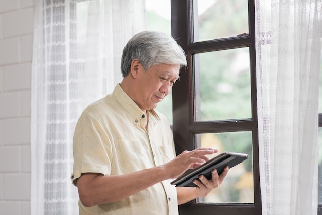 Homme âgé Asiatique à L'aide De Tablette, Vérification Des Médias Sociaux Près De La Fenêtre Dans Le Salon à La Maison. Mode De Vie Senior Hommes à La Maison Concept.
