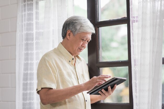 Homme âgé asiatique à l&#39;aide de tablette, vérification des médias sociaux près de la fenêtre dans le salon à la maison. Mode de vie senior hommes à la maison concept.