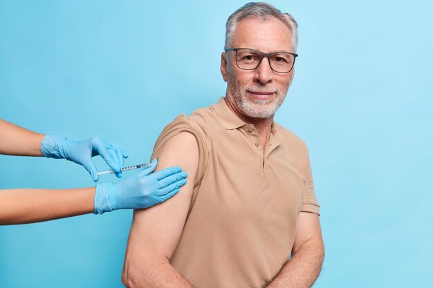 Un homme âgé aux cheveux gris barbu se fait vacciner contre le coronavirus se protège du virus porte des lunettes et un t-shirt a l'air déterminé isolé sur un mur bleu