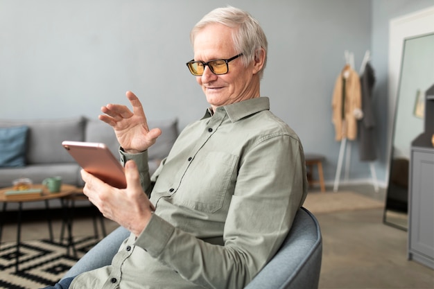 Homme âgé ayant un appel vidéo à l'aide d'une tablette assis sur une chaise dans le salon