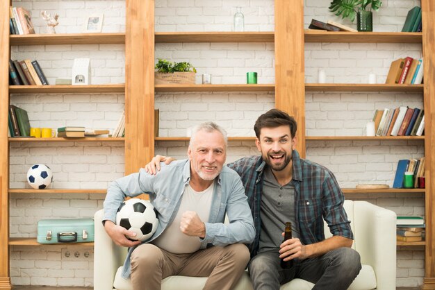 Homme âgé avec ballon et jeune homme avec une bouteille devant la télé sur un canapé
