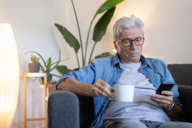 Homme âgé buvant du café dans le salon et utilisant le téléphone. Homme mûr paisible dans des vêtements décontractés assis dans un fauteuil, regardant l'écran mobile, lisant la newsletter. Matin, concept d'appareils numériques