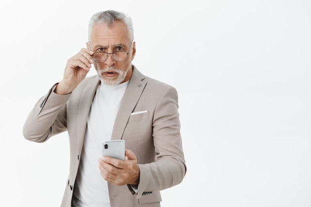 Homme âgé confus en costume tenant un téléphone mobile et à la recherche