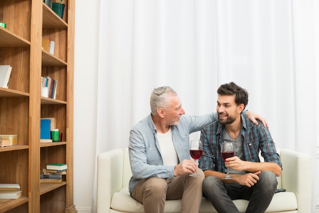 Homme âgé étreignant jeune homme souriant avec des verres de vin sur le canapé dans la chambre
