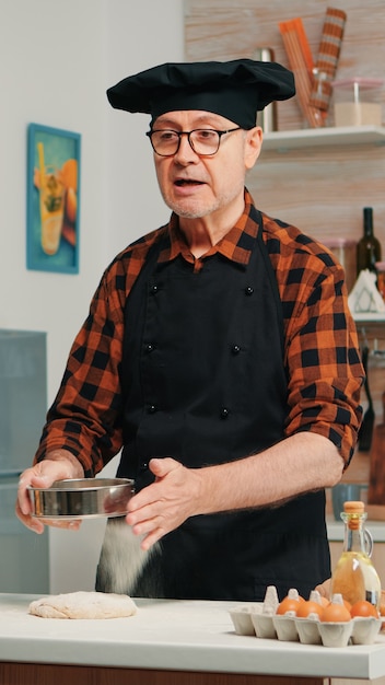 Un homme âgé gai enregistrant la recette étape par étape dans la cuisine de la houe. Chef influenceur blogueur à la retraite utilisant la technologie Internet pour communiquer, bloguer sur les réseaux sociaux avec un équipement numérique
