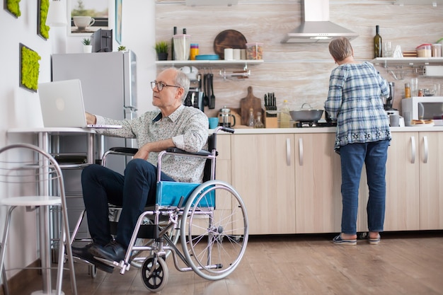 Homme âgé handicapé en fauteuil roulant travaillant sur un ordinateur portable dans la cuisine pendant que sa femme prépare un délicieux petit-déjeuner pour tous les deux. Homme utilisant la technologie moderne tout en travaillant à domicile.