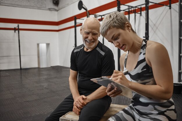 Homme âgé heureux charismatique avec un corps athlétique musclé assis au centre de remise en forme avec une jeune entraîneuse blonde qui écrit les résultats après un entraînement personnel avec son client senior