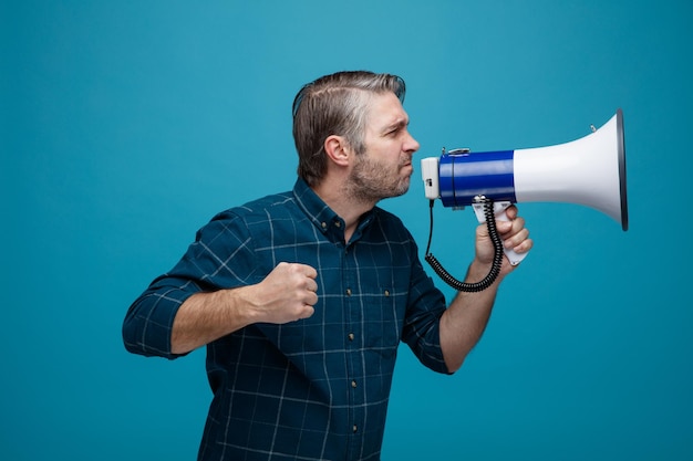 Homme d'âge moyen aux cheveux gris en chemise de couleur foncée criant dans un mégaphone étant excité serrant le poing debout sur fond bleu