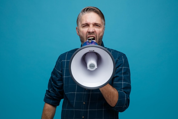 Homme d'âge moyen aux cheveux gris en chemise de couleur foncée criant dans un mégaphone avec une expression agressive debout sur fond bleu