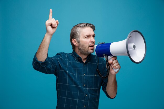 Homme d'âge moyen aux cheveux gris en chemise de couleur foncée criant dans un mégaphone pointant avec l'index debout sur fond bleu