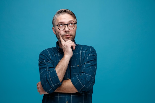 Homme d'âge moyen aux cheveux gris en chemise de couleur foncée portant des lunettes levant les yeux perplexe debout sur fond bleu