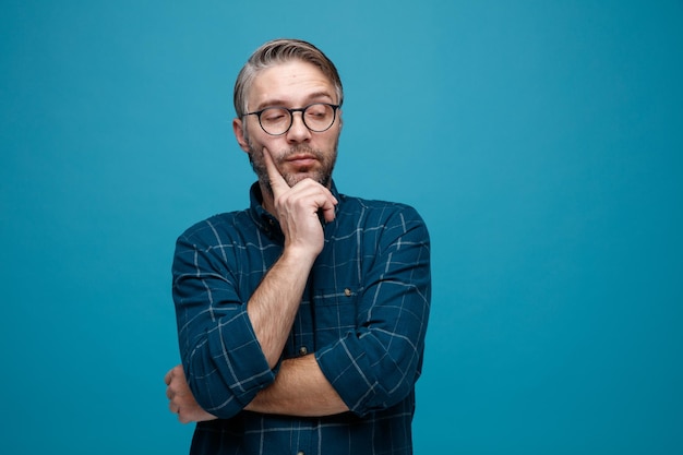 Homme d'âge moyen aux cheveux gris en chemise de couleur foncée portant des lunettes regardant de côté avec une expression sceptique pensant debout sur fond bleu