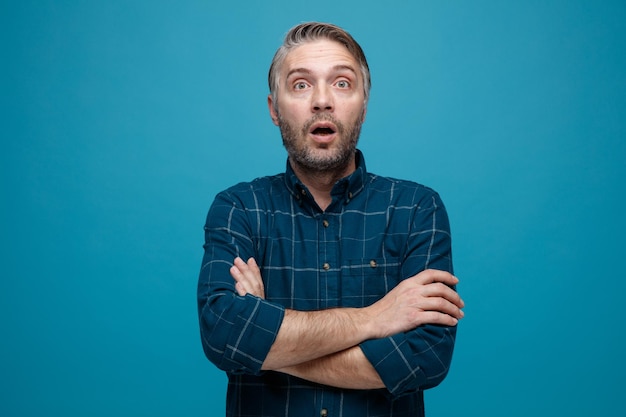 Homme d'âge moyen aux cheveux gris en chemise de couleur foncée regardant la caméra avec les bras croisés sur sa poitrine étant étonné et surpris debout sur fond bleu