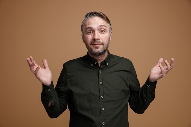 Homme d'âge moyen aux cheveux gris en chemise de couleur foncée regardant la caméra confus levant les bras ayant des doutes debout sur fond marron