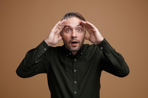 Homme d'âge moyen aux cheveux gris en chemise de couleur foncée regardant la caméra ouvrir les yeux étonnés et choqués debout sur fond marron