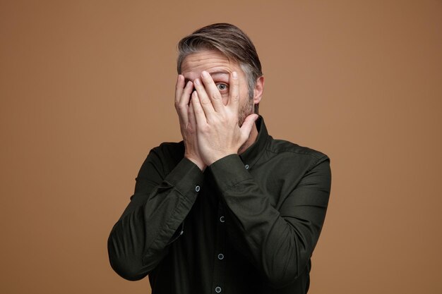 Homme d'âge moyen aux cheveux gris en chemise de couleur foncée regardant la caméra à travers les paumes effrayé et inquiet debout sur fond marron