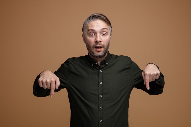 Homme d'âge moyen aux cheveux gris en chemise de couleur foncée regardant vers le bas étant excité pointant avec l'index vers le bas debout sur fond marron