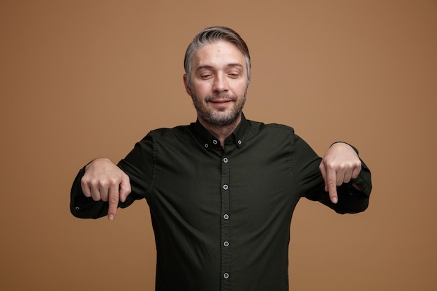 Homme d'âge moyen aux cheveux gris en chemise de couleur foncée regardant vers le bas heureux et positif pointant avec l'index vers le bas debout sur fond marron