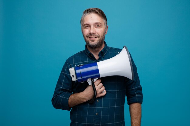 Homme d'âge moyen aux cheveux gris en chemise de couleur foncée tenant un mégaphone regardant la caméra heureux et positif souriant confiant debout sur fond bleu