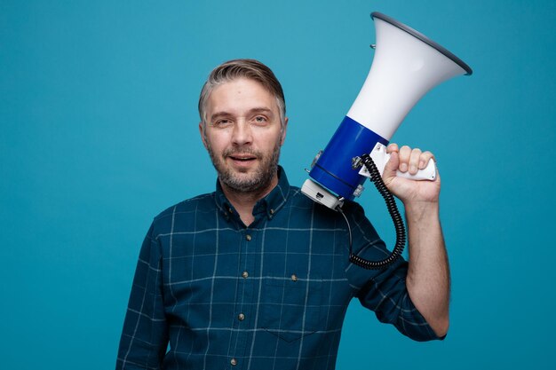Homme d'âge moyen aux cheveux gris en chemise de couleur foncée tenant un mégaphone regardant la caméra souriant confiant debout sur fond bleu