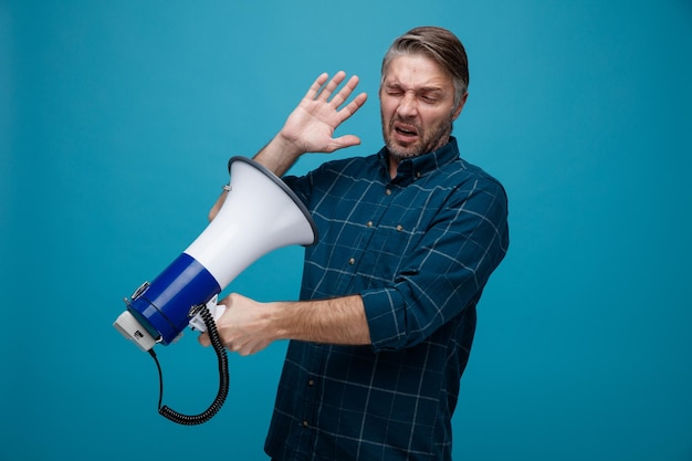 Homme d'âge moyen aux cheveux gris en chemise de couleur foncée tenant un mégaphone en le regardant être mécontent debout sur fond bleu