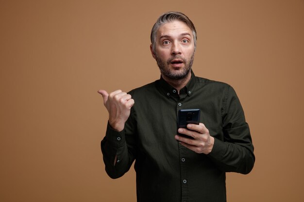homme d'âge moyen aux cheveux gris en chemise de couleur foncée tenant un smartphone pointant avec le pouce sur le côté à l'air étonné et surpris debout sur fond marron