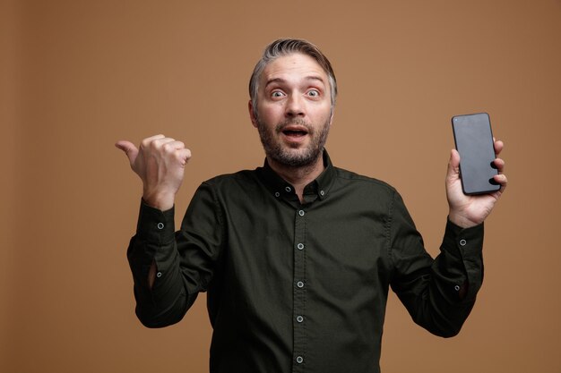 Homme d'âge moyen aux cheveux gris en chemise de couleur foncée tenant un smartphone pointant avec le pouce sur le côté heureux et surpris en regardant la caméra debout sur fond marron