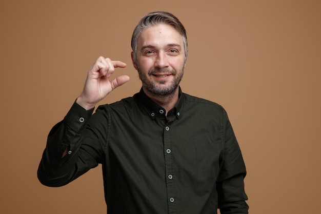 Homme d'âge moyen aux cheveux gris en chemise vert foncé regardant la caméra en souriant montrant un signe de petite taille avec les doigts debout sur fond marron