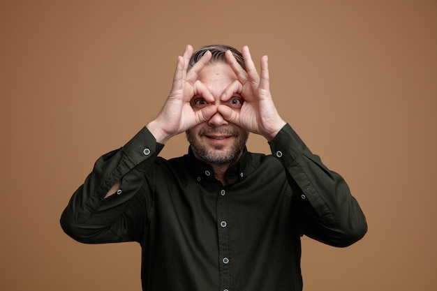 homme d'âge moyen aux cheveux gris en chemise vert foncé regardant la caméra à travers les doigts faisant un geste binoculaire souriant debout sur fond marron