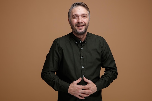 Homme d'âge moyen aux cheveux gris en chemise vert foncé souriant joyeusement main dans la main regardant la caméra debout sur fond marron