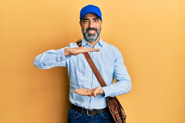 Homme d'âge moyen avec barbe et cheveux gris portant une casquette de courrier de livraison gesticulant avec les mains montrant un signe de grande et grande taille, symbole de mesure. souriant en regardant la caméra. notion de mesure.