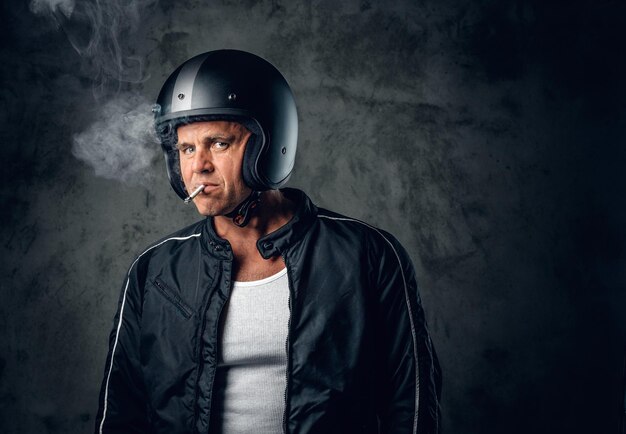 Homme d'âge moyen en casque de moto et veste en cuir fumant une cigarette sur fond gris.
