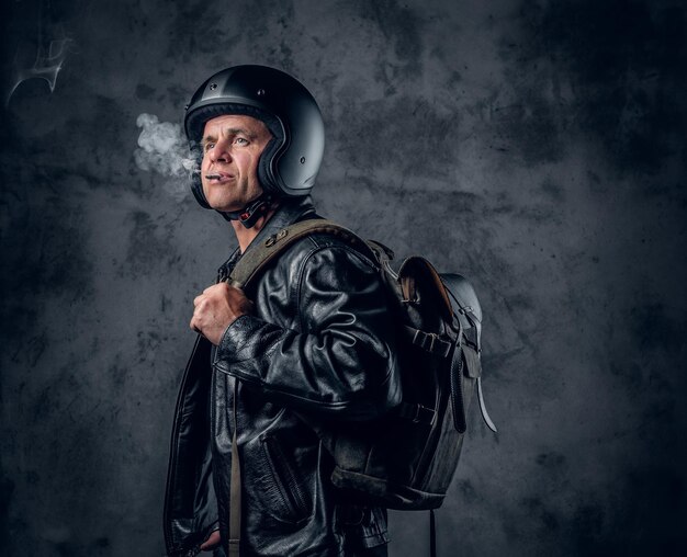 Homme d'âge moyen en casque de moto et veste en cuir avec sac à dos fumant une cigarette sur fond gris.
