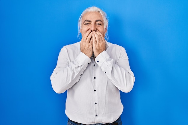 Photo gratuite homme d'âge moyen avec des cheveux gris debout sur fond bleu riant et rire embarrassé couvrant la bouche avec les mains concept de potins et de scandale