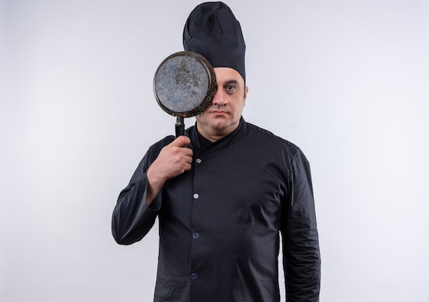 Homme d'âge moyen cuisinier en uniforme de chef couvert oeil avec poêle à frire sur mur blanc isolé