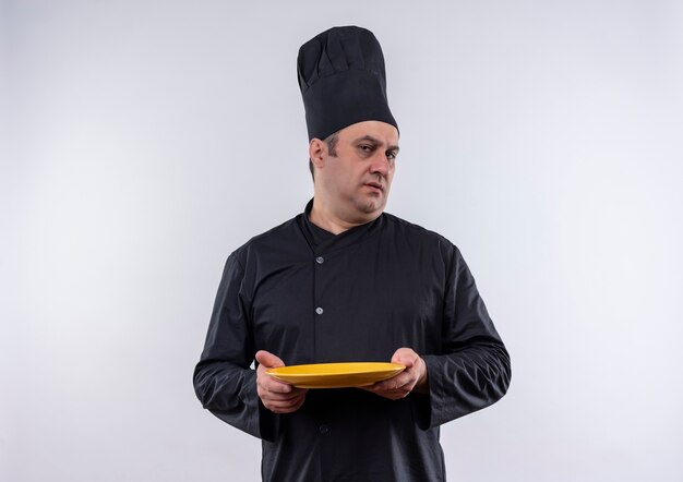 Homme d'âge moyen cuisinier en uniforme de chef tenant la plaque sur un mur blanc isolé